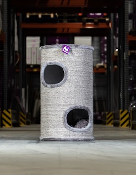 A tall, cylindrical cat tower with two openings is placed in an industrial or warehouse setting. It has a textured, grey exterior with a small purple tag bearing a logo or design.