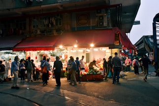 A bustling marketplace scene with a small shop illuminated by bright lights. Numerous people are shopping and socializing under a red awning. Vegetables and other goods are displayed, attracting a diverse crowd.