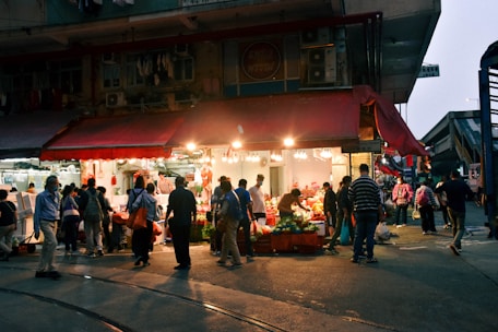 A bustling marketplace scene with a small shop illuminated by bright lights. Numerous people are shopping and socializing under a red awning. Vegetables and other goods are displayed, attracting a diverse crowd.