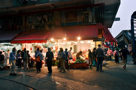 A bustling marketplace scene with a small shop illuminated by bright lights. Numerous people are shopping and socializing under a red awning. Vegetables and other goods are displayed, attracting a diverse crowd.