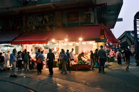 A bustling marketplace scene with a small shop illuminated by bright lights. Numerous people are shopping and socializing under a red awning. Vegetables and other goods are displayed, attracting a diverse crowd.