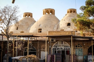 Three large, dome-shaped structures with small openings are set atop a building resembling a traditional bathhouse or bazaar. Arched windows and brickwork enhance the architectural details. Surrounding the structures are trees, and fabric or rugs are displayed in front of the building, suggesting a marketplace scene.