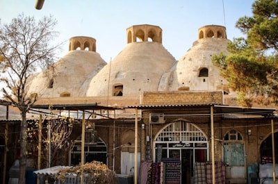 Three large, dome-shaped structures with small openings are set atop a building resembling a traditional bathhouse or bazaar. Arched windows and brickwork enhance the architectural details. Surrounding the structures are trees, and fabric or rugs are displayed in front of the building, suggesting a marketplace scene.