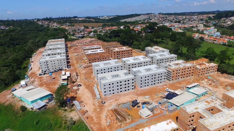 The image shows an aerial view of a construction site with several multi-story apartment buildings at different stages of completion. The buildings are arranged in rows with some fully constructed and others partially built. The surroundings include green areas and a residential neighborhood in the distance.