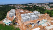 The image shows an aerial view of a construction site with several multi-story apartment buildings at different stages of completion. The buildings are arranged in rows with some fully constructed and others partially built. The surroundings include green areas and a residential neighborhood in the distance.