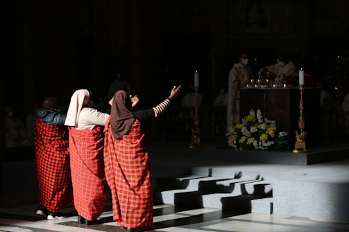 Three individuals dressed in traditional red and black checkered garments are standing with their backs to the camera, facing a church altar. The scene is dimly lit, highlighting the figures and the altar. There are priests in white robes near the altar, which is adorned with candles and a flower arrangement.