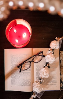 An open book with pale yellow pages, lying next to a pair of reading glasses and a flickering candle.