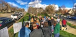 A group of people are gathered outdoors in a suburban neighborhood near several parked cars. They appear to be looking at some printed materials or diagrams displayed on stands. The environment is sunny with green lawns and a backdrop of trees and houses lining the road.