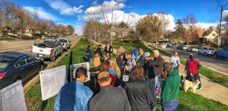 A group of people are gathered outdoors in a suburban neighborhood near several parked cars. They appear to be looking at some printed materials or diagrams displayed on stands. The environment is sunny with green lawns and a backdrop of trees and houses lining the road.