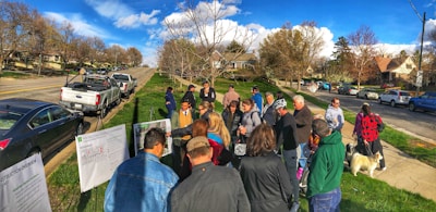 A group of people are gathered outdoors in a suburban neighborhood near several parked cars. They appear to be looking at some printed materials or diagrams displayed on stands. The environment is sunny with green lawns and a backdrop of trees and houses lining the road.