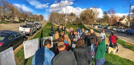 A group of people are gathered outdoors in a suburban neighborhood near several parked cars. They appear to be looking at some printed materials or diagrams displayed on stands. The environment is sunny with green lawns and a backdrop of trees and houses lining the road.