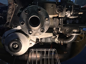 A close-up view of a mechanical engine component, featuring a central circular metallic part with bolts and surrounding machinery. Various pipes and wires are visible, indicating a complex assembly. The lighting highlights the metallic and industrial aspects, providing a detailed look at the pistons and gears.