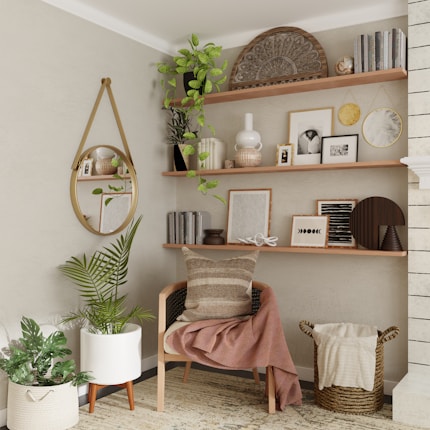 A cozy living space corner features a wooden chair with a striped cushion and a folded throw blanket. Above the chair, shelves are adorned with various decorative items including potted plants, books, framed photographs, and abstract art pieces. On the wall hangs a round mirror reflecting part of the room. A large green plant and a woven basket containing a blanket sit on the floor, contributing to the warm, inviting atmosphere.