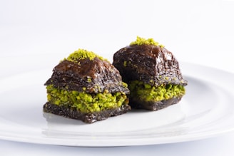 Two pieces of dessert are displayed on a white plate. The dessert appears to be baklava, made with layers of flaky pastry and a filling that includes vibrant green pistachios. The pastry is coated with a dark, shiny syrup, giving it a rich and glossy appearance.
