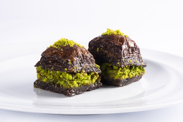 Two pieces of dessert are displayed on a white plate. The dessert appears to be baklava, made with layers of flaky pastry and a filling that includes vibrant green pistachios. The pastry is coated with a dark, shiny syrup, giving it a rich and glossy appearance.