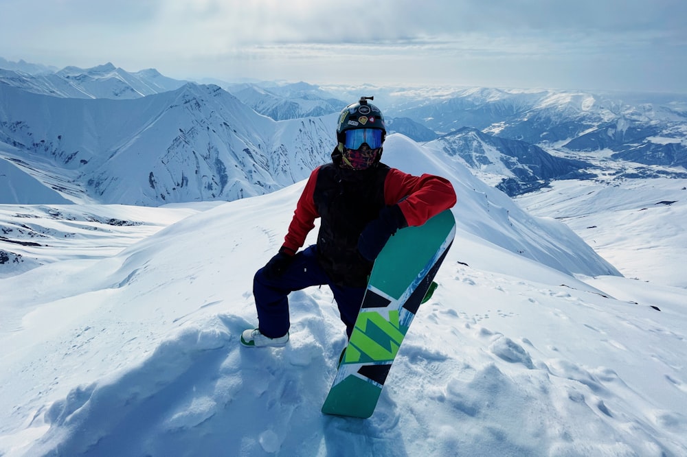 man in black jacket and blue pants sitting on green and black snowboard on snow covered