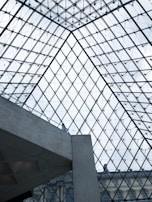 A glass and metal geometric structure forms the architectural ceiling, composed of numerous triangular panes. Below, a concrete element intersects the view, with a person standing and observing the surroundings. Structural lines create a symmetrical pattern, offering a glimpse of a classic building façade under the glass.