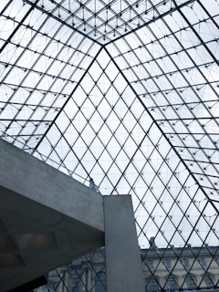 A glass and metal geometric structure forms the architectural ceiling, composed of numerous triangular panes. Below, a concrete element intersects the view, with a person standing and observing the surroundings. Structural lines create a symmetrical pattern, offering a glimpse of a classic building façade under the glass.