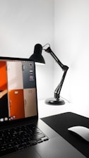 A minimalist workspace featuring a sleek black desk lamp and an open laptop displaying a desktop background with colorful lockers. The setting is brightly lit, highlighting the clean and modern aesthetic. A wireless mouse is also visible on a dark mouse pad.