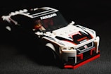 A model car made from interlocking plastic bricks, resembling a racing vehicle. The car is prominently white with black and red detailing, and features brand logos such as Nissan and Nismo. The image is sharply focused on the car, with a dark, shadowy background that enhances the vibrant colors of the model.