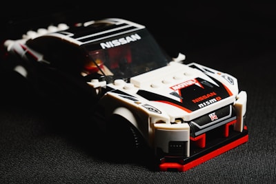 A model car made from interlocking plastic bricks, resembling a racing vehicle. The car is prominently white with black and red detailing, and features brand logos such as Nissan and Nismo. The image is sharply focused on the car, with a dark, shadowy background that enhances the vibrant colors of the model.
