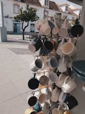 A display rack features an array of ceramic mugs in various colors and designs hanging in an outdoor setting. The background includes a cobblestone path, a bench, a tree, and a white building with traditional architecture. The scene is sunlit, casting shadows on the ground.