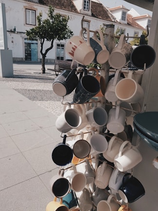 A display rack features an array of ceramic mugs in various colors and designs hanging in an outdoor setting. The background includes a cobblestone path, a bench, a tree, and a white building with traditional architecture. The scene is sunlit, casting shadows on the ground.