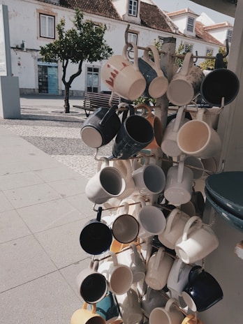 A display rack features an array of ceramic mugs in various colors and designs hanging in an outdoor setting. The background includes a cobblestone path, a bench, a tree, and a white building with traditional architecture. The scene is sunlit, casting shadows on the ground.