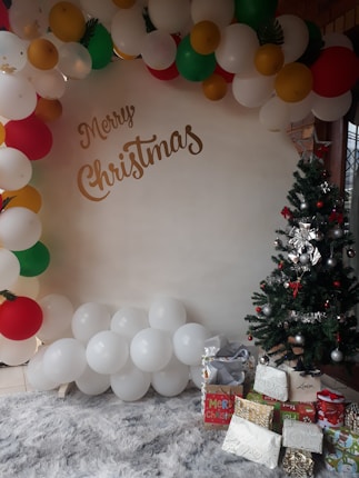 A festive scene featuring a cluster of balloons in red, white, green, and gold hues arranged in an arch pattern. Below the balloons, several wrapped gifts are placed beside a decorated Christmas tree adorned with silver and red ornaments. The phrase 'Merry Christmas' is written in elegant script on a backdrop. The area is carpeted with a fluffy, light-colored rug.