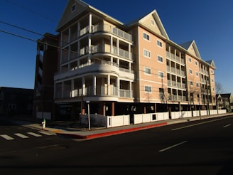 A multi-story residential building with a light brown and white facade, featuring multiple balconies and a sloped roof. It is set on a corner lot with white fencing along the perimeter. The surrounding area includes a road with a crosswalk and a fire hydrant. The scene is captured during daylight with clear blue skies.