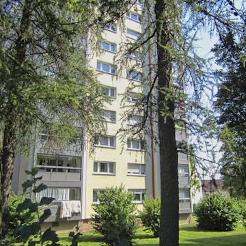 A multi-story residential building with several windows and balconies is partially obscured by tall trees and lush green foliage. White fabrics appear to be drying on one of the balconies, which suggests domestic life. Sunlight illuminates the building, creating a warm and welcoming atmosphere.