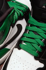A close-up view of athletic shoes featuring vibrant green laces, black detailing, and white leather accents. The image shows a partial side and top view of the shoes, emphasizing their distinct color contrast and texture.