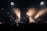 A rock band performs energetically on stage, with members playing guitars and singing into microphones. Bright spotlights create dramatic lighting effects, highlighting the performers against a dark background. The ambiance is lively, with audience heads subtly visible in the foreground.