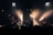A rock band performs energetically on stage, with members playing guitars and singing into microphones. Bright spotlights create dramatic lighting effects, highlighting the performers against a dark background. The ambiance is lively, with audience heads subtly visible in the foreground.