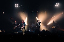 A rock band performs energetically on stage, with members playing guitars and singing into microphones. Bright spotlights create dramatic lighting effects, highlighting the performers against a dark background. The ambiance is lively, with audience heads subtly visible in the foreground.