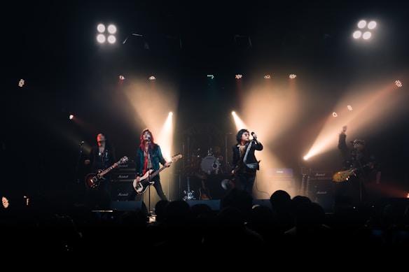 A rock band performs energetically on stage, with members playing guitars and singing into microphones. Bright spotlights create dramatic lighting effects, highlighting the performers against a dark background. The ambiance is lively, with audience heads subtly visible in the foreground.
