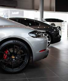 A showroom displays two luxury cars parked side by side. Both cars are sleek and modern, with the car in the foreground being silver and featuring a prominent wheel with a red brake caliper. The second car in the background is black.