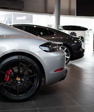 A showroom displays two luxury cars parked side by side. Both cars are sleek and modern, with the car in the foreground being silver and featuring a prominent wheel with a red brake caliper. The second car in the background is black.
