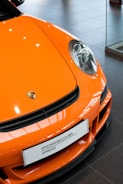 An orange sports car featuring a prominent Porsche emblem on the hood sits in a showroom. The sleek and polished exterior is highlighted under bright lighting, emphasizing the car's aerodynamic design and front headlights.
