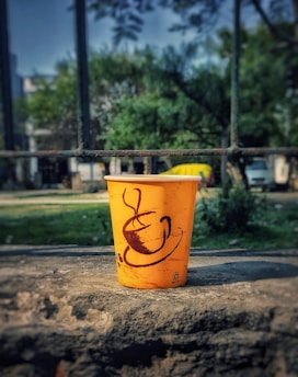 A bright orange disposable cup with a coffee cup illustration is placed on a rough stone surface. The cup is in focus with a background of greenery and blurred structures.