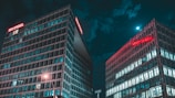 Two modern office buildings with glass facades, illuminated by streetlights and the moon at night. The logo for 'Der Spiegel' is visible in bright red neon on the top of one building. The scene has a teal color tone, with a clear night sky partially covered by clouds.