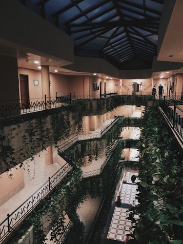 A multi-story indoor atrium features several floors with railings adorned with lush green hanging plants. The ceiling is made of glass panels, allowing natural light to filter through. There are warm lights illuminating the walls, and a checkered floor pattern is visible on the ground level. Two people are seen walking along the top floor.