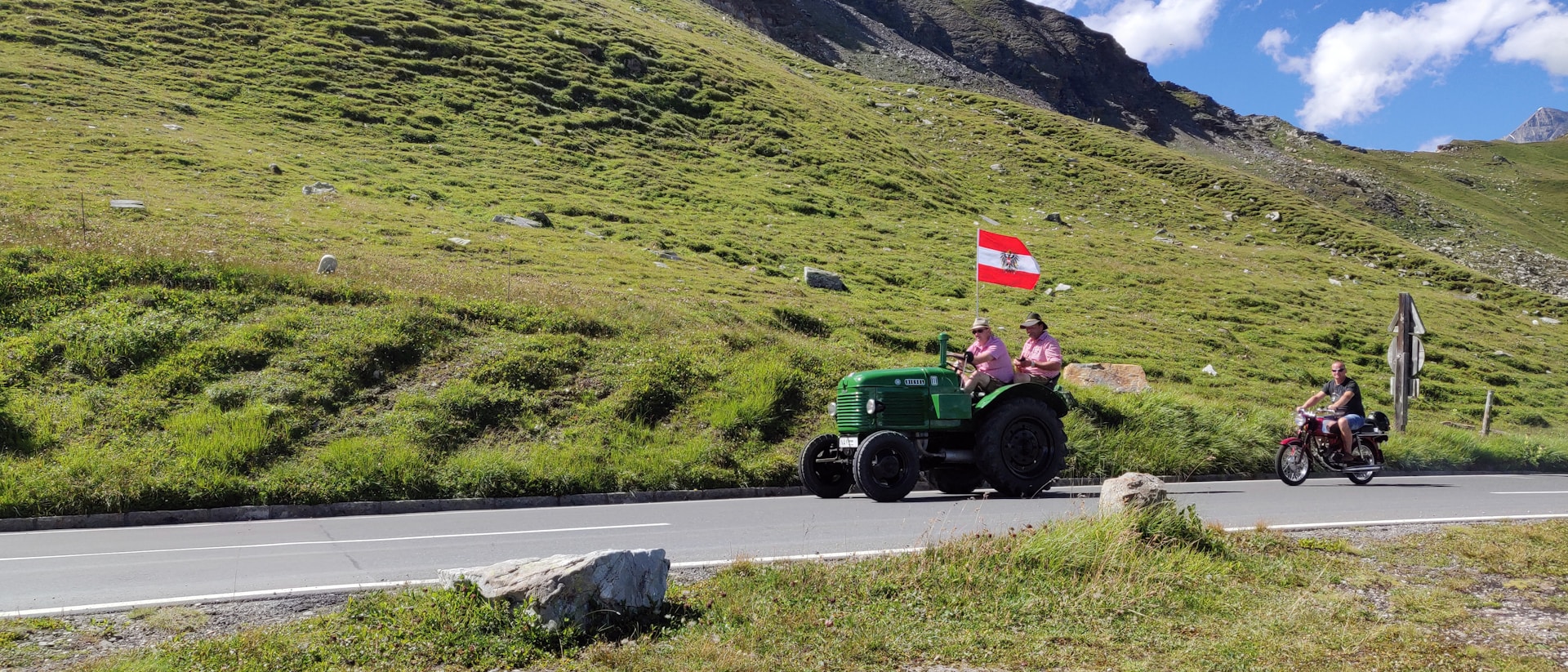 A rural scene features two individuals on a green tractor carrying a flag with red and white stripes and a black emblem. Next to them, another person rides a motorbike on a road that winds through a vibrant green grassy landscape, set against a backdrop of hills and a bright blue sky with scattered clouds.