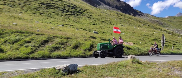 A rural scene features two individuals on a green tractor carrying a flag with red and white stripes and a black emblem. Next to them, another person rides a motorbike on a road that winds through a vibrant green grassy landscape, set against a backdrop of hills and a bright blue sky with scattered clouds.