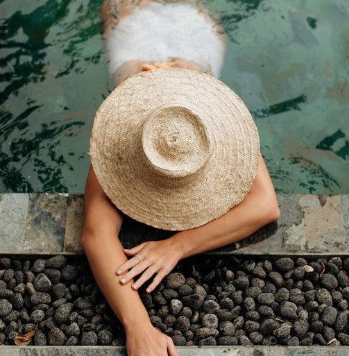 person wearing brown straw hat sitting on black stones near body of water during daytime