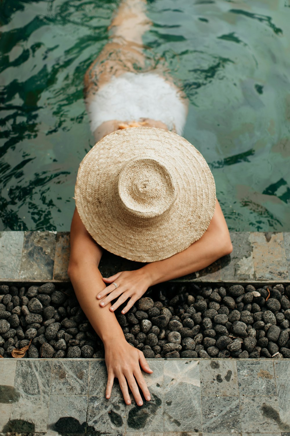 personne portant un chapeau de paille brun assise sur des pierres noires près d’un plan d’eau pendant la journée