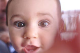 A close-up of a young child's face with wide, curious eyes and a slightly open mouth. The focus is primarily on the eyes, giving a sense of innocence and wonder. A hand partially covers the right side of the image, creating a blurred effect.