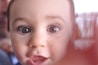 A close-up of a young child's face with wide, curious eyes and a slightly open mouth. The focus is primarily on the eyes, giving a sense of innocence and wonder. A hand partially covers the right side of the image, creating a blurred effect.