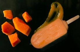 Several pieces of ripe orange mango are arranged alongside a melting mango popsicle on a dark surface. The popsicle features a visible splash of green and orange liquid, suggesting freshness and indulgence.