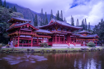 A traditional Japanese-style building is situated by a tranquil pond, surrounded by lush greenery and tall, slender trees. The structure is painted in a striking red color with intricate architectural details, including curved roofs and ornate decorations. The serene setting is complemented by the presence of mountains in the background and a gentle fountain in the pond.